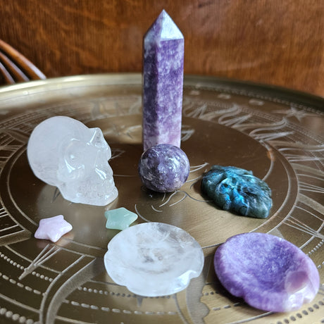 A selection of crystals on a metal surface, including a Lepidolite Generator, lepidolite sphere, a clear quartz crystal skull, thumbstones, crystal stars and a viking carving