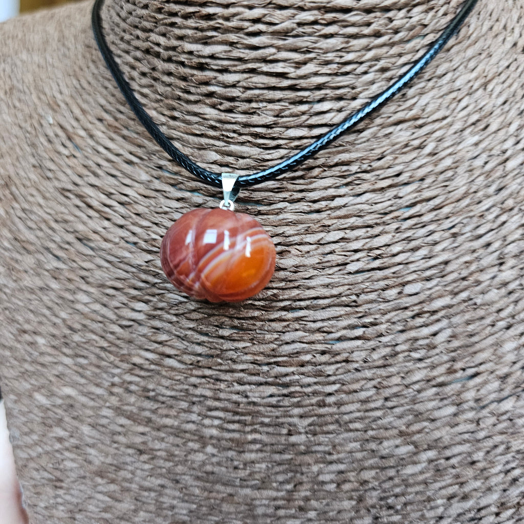 A pumpkin-shaped carnelian crystal pendant attached to a black cord necklace, displayed on a textured background
