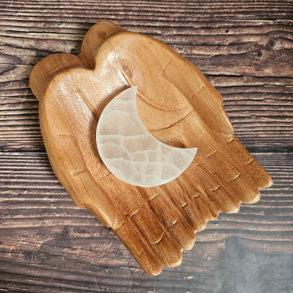 A selenite crystal charging plate shaped as a crescent moon, placed on a wooden pair of hands