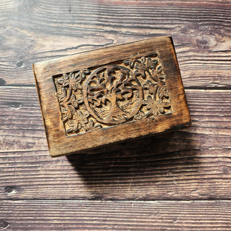 A wooden box with a carved Tree of Life design on the lid, placed on a wooden surface.