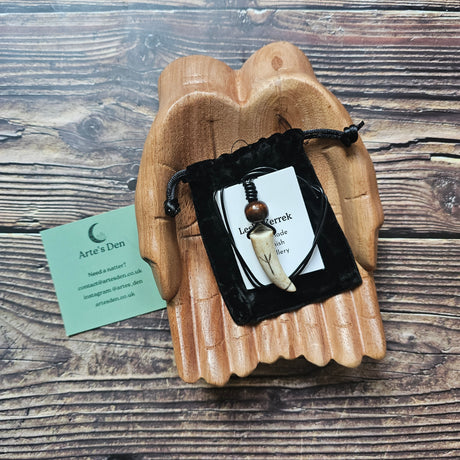 A handcarved antler pendant necklace with a black cord, featuring the Algiz Rune, placed on a wooden surface with a business card beside it.