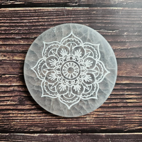 A round selenite charging plate with a mandala pattern etched into it, placed on a wooden surface.