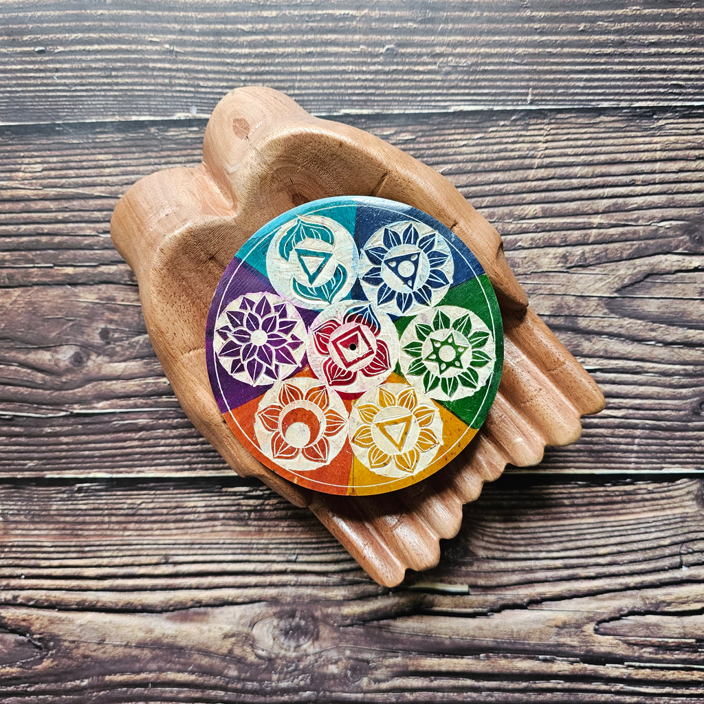 A colourful soapstone incense holder with chakra symbols, placed on a wooden pair of hands on a wooden surface. 