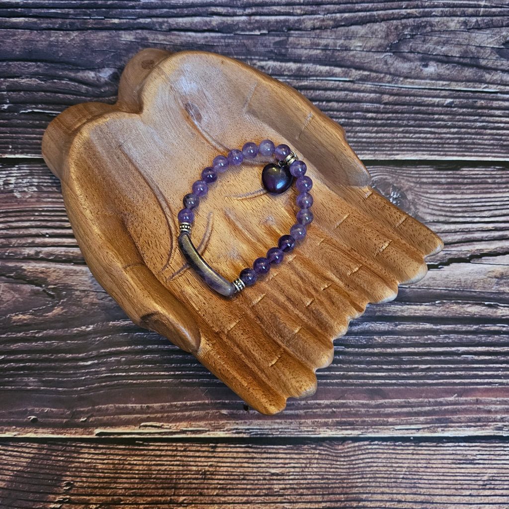 Wooden hands holding a purple amethyst bead bracelet with a heart-shaped bead and bar quartz accents.