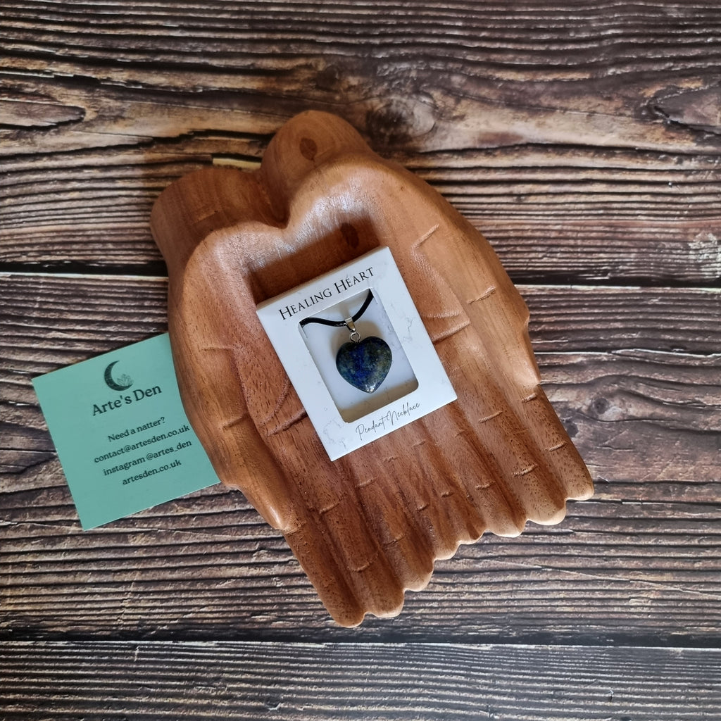 A blue crystal heart on a black cord necklace inside a white display box, displayed on a pair of wooden hands