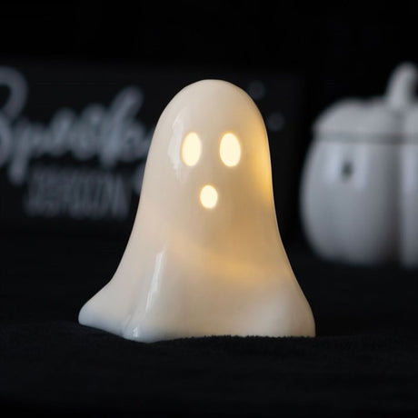 A ceramic ghost-shaped LED light with a visible LED light source, placed on a dark surface next to a white pumpkin 