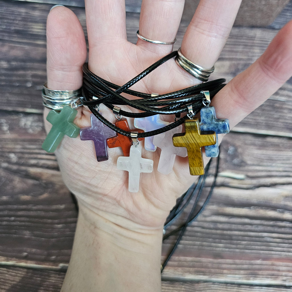 Several cross-shaped crystal pendants with a black cord necklace held in someone's hand. 