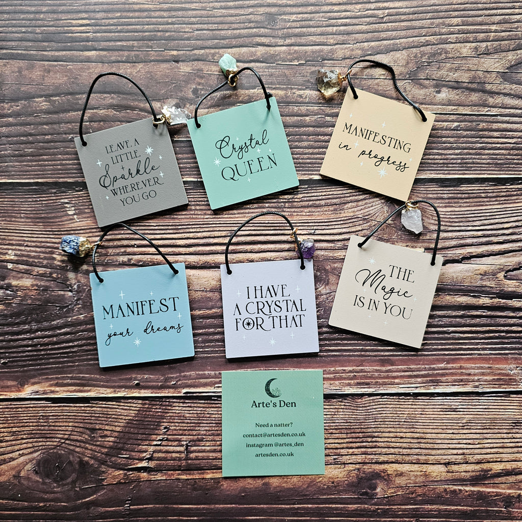 A selection of mini signs with attached crystal charms, each inscribed with different phrases, displayed on a wooden surface.