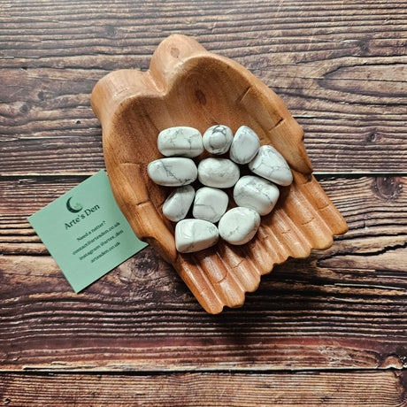 Several white howlite tumbled crystals placed in a pair of wooden hands on a wooden surface next to a green Arte's Den business card 