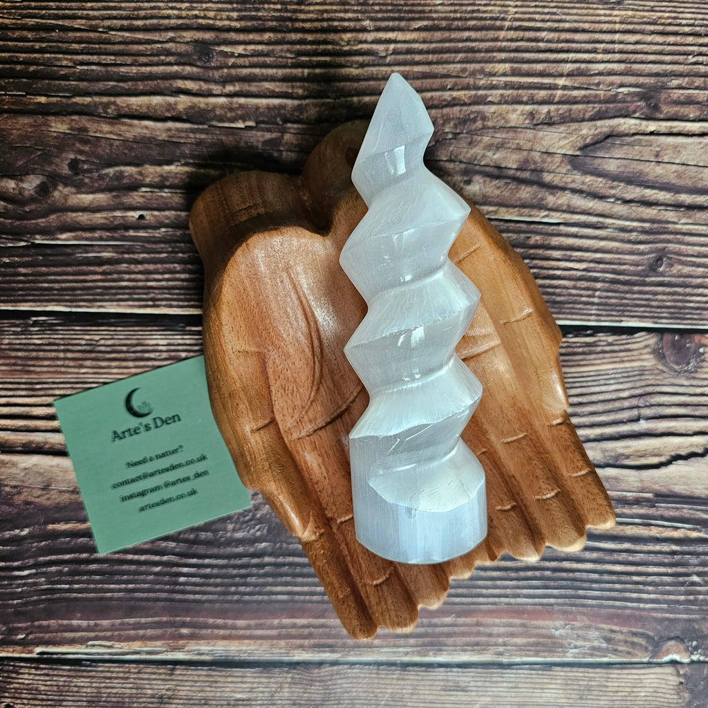A selenite "unicorn horn" which is a spiral tower, laid on a wooden pair of hands on a wooden background