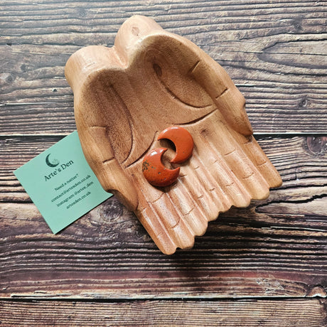 Two red jasper crescent moon shaped crystals placed on a pair of wooden hands on a wooden surface, with a business card labelled 'Arte's Den'.