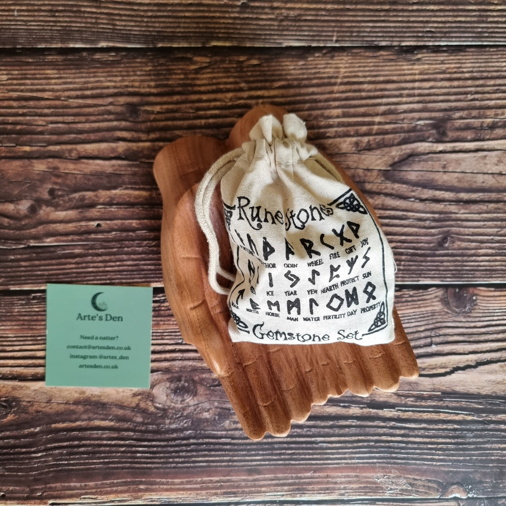 A beige "rune stones" bag adorned with various black runes, displayed in a pair of wooden hands, on a wooden surface with a business card labelled 'Arte's Den' on the side.