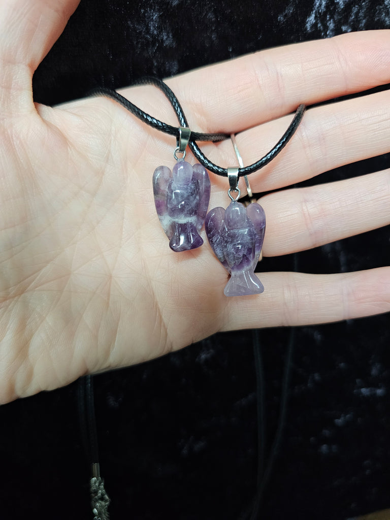 Two angel-shaped crystal pendant attached to black cord necklaces held in a person's hand on a dark background