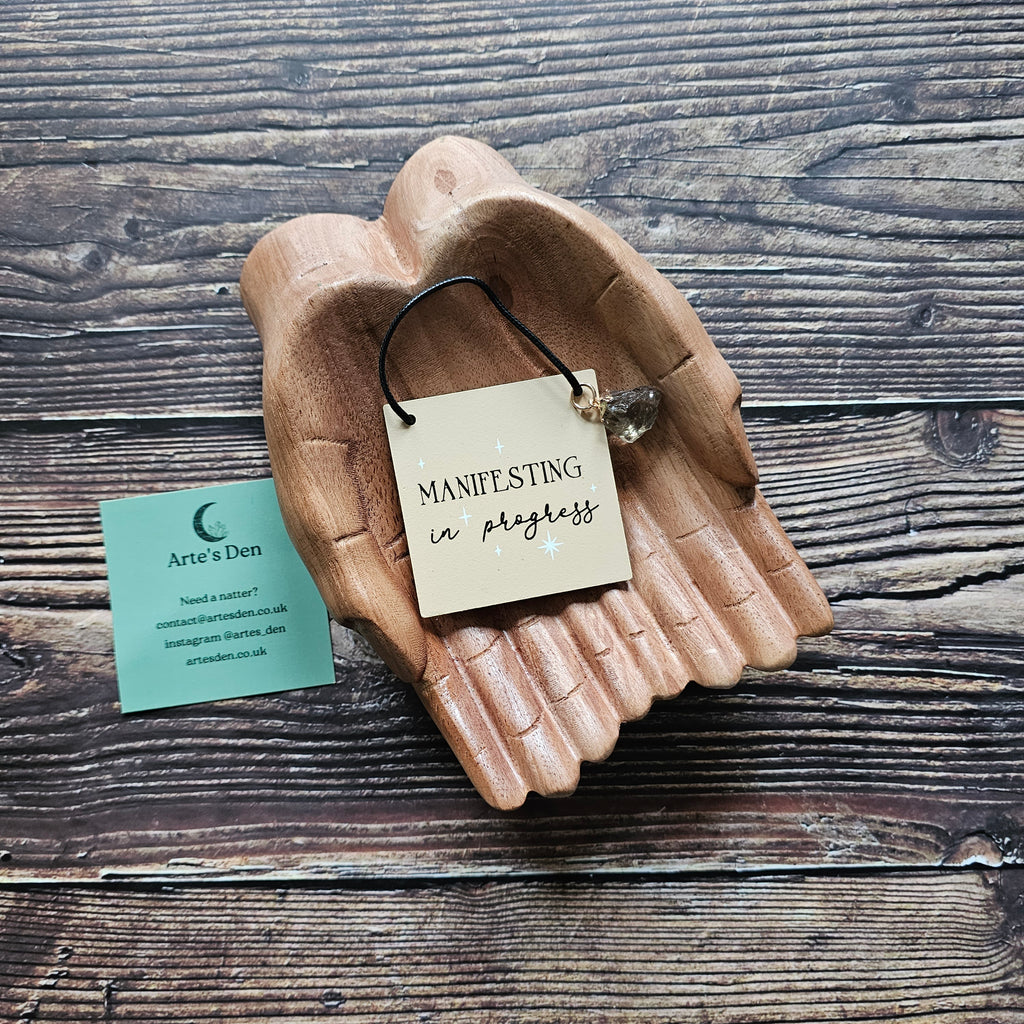A mini sign with attached crystal charm, inscribed with the phrase "Manifesting in Progress" displayed on a pair of wooden hands on a wooden surface. 