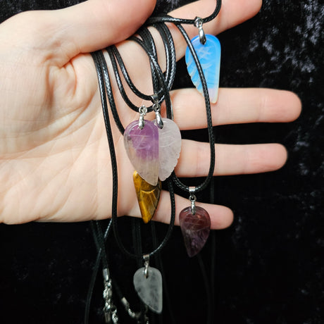 A collection of angel wing-shaped crystal pendants in various colours, attached to black cord necklaces, held in a person's hand.