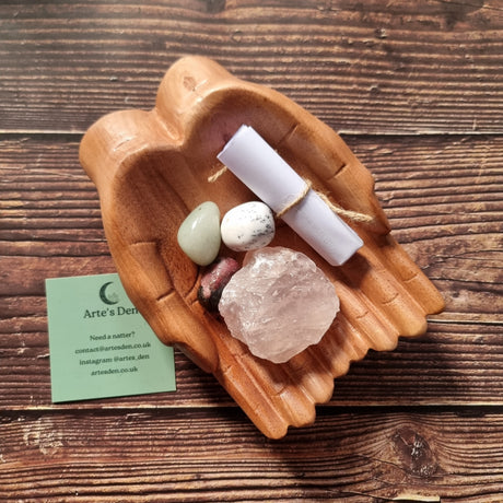A collection of crystals and a scroll of paper displayed on a pair of wooden hands, with a business card that reads 'Arte's Den'.