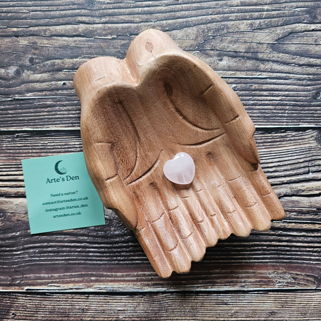 A rose quartz crystal heart stone placed on a wooden pair of hands.