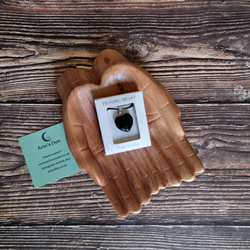 A black crystal heart on a black cord necklace inside a white display box, displayed on a pair of wooden hands