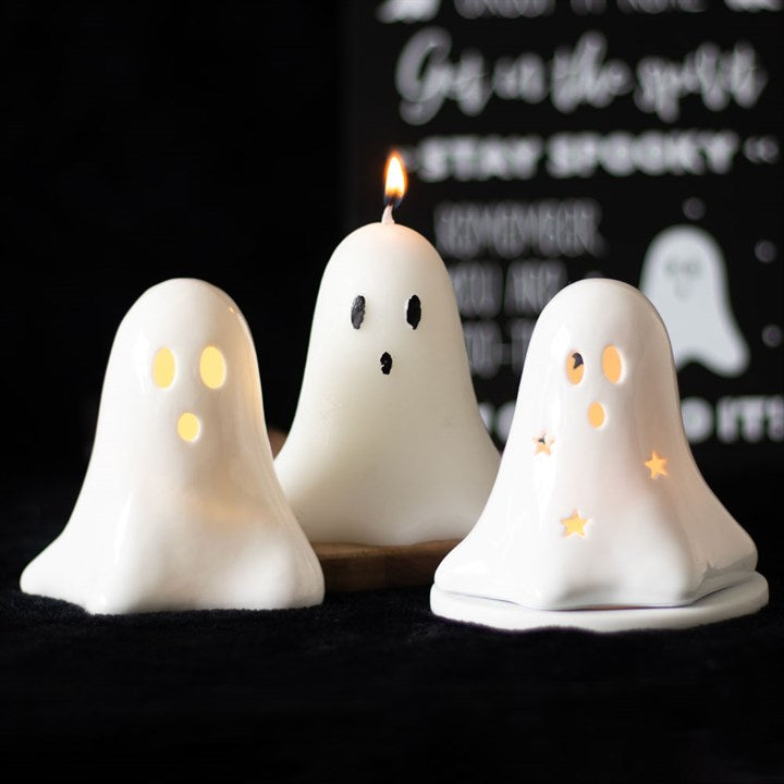 A ceramic ghost-shaped LED light with a visible LED light source, placed next to a ghost shaped lit candle and a ghost shaped tealight holder