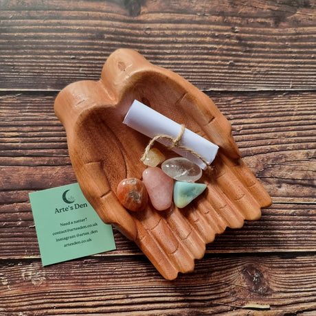 A collection of polished crystals and a scroll of paper displayed in a pair of wooden hands, with a business card that reads 'Arte's Den'.