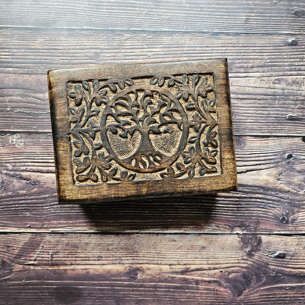 A wooden box with a carved Tree of Life design on the lid, placed on a wooden surface.