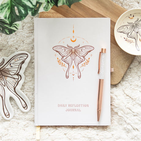 A white lined notebook with a unique luna moth design on the cover, accompanied by a clear quartz pen, placed on a wooden surface with a few decorative items around.