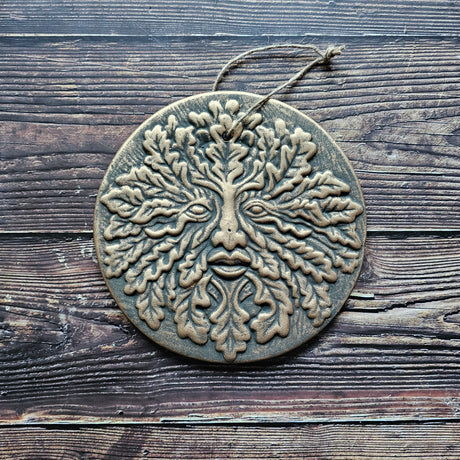 A round terracotta plaque with a bronze effect finish, featuring the Green Man symbol with leaf patterns for eyes and branches for a nose and mouth, mounted on a twine loop.