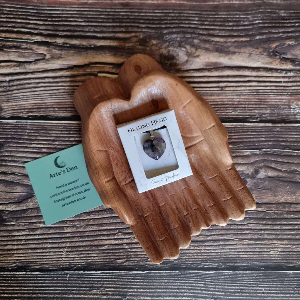 A purple crystal heart on a black cord necklace inside a white display box, displayed on a pair of wooden hands