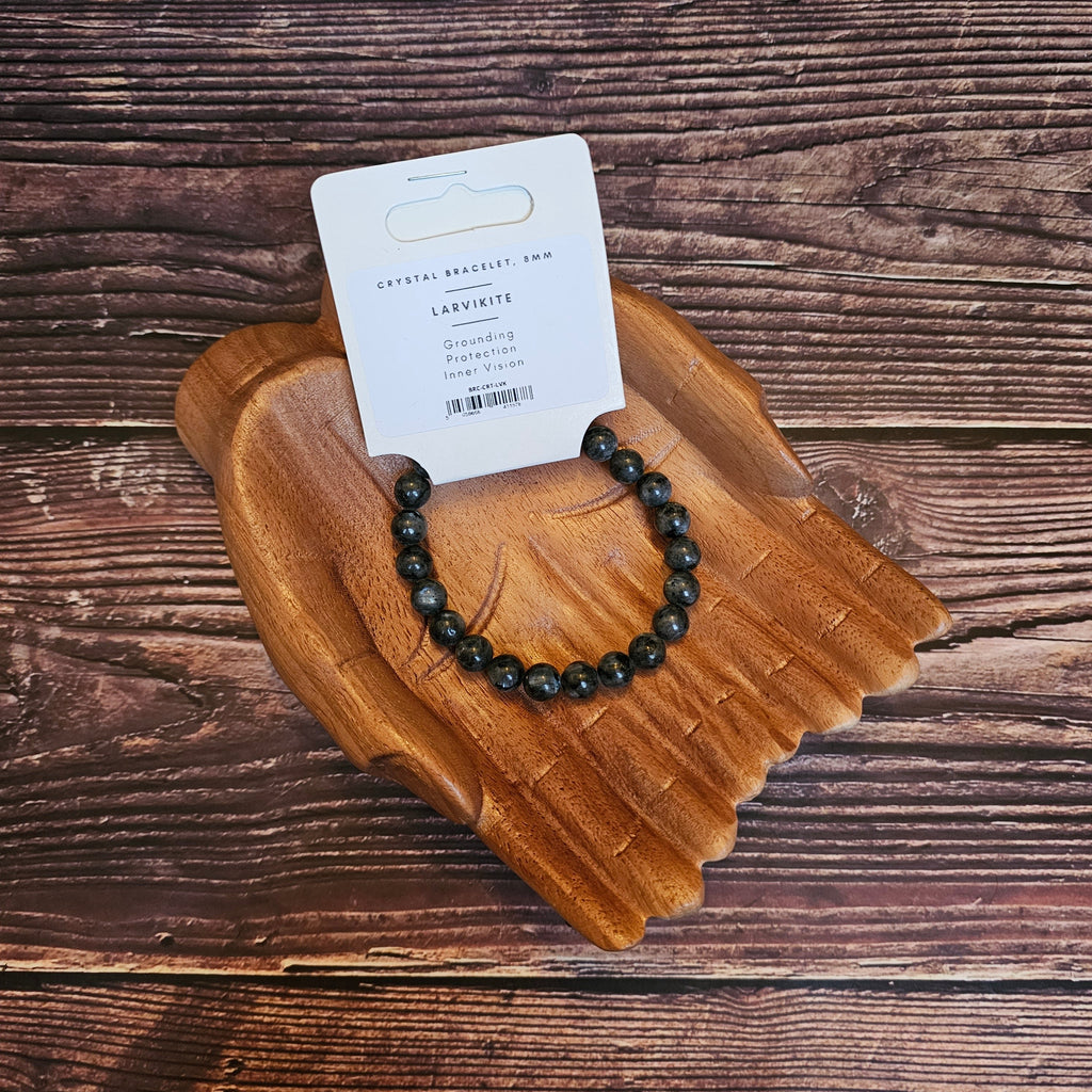A beaded bracelet with black and gray stones, displayed on a wooden bracelet holder with a card in the background indicating the product is called 'Larvikite Beaded Bracelet' 