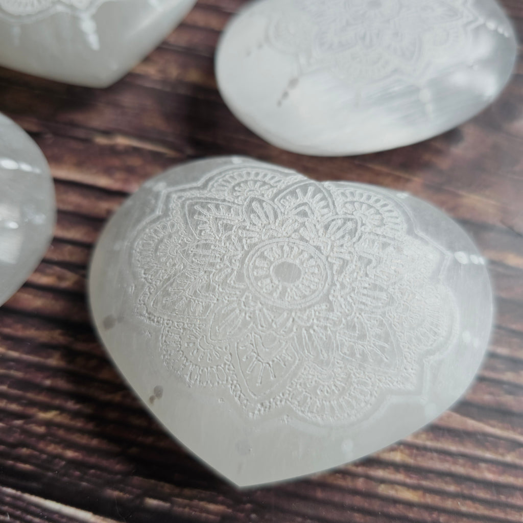A Selenite heart, carved with intricate mandala design, laid on a wooden surface
