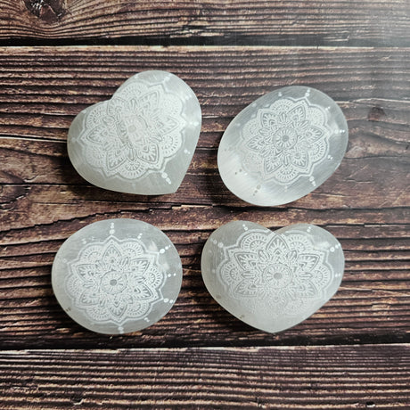 Four Selenite stones, two shaped as hearts, two as ovals, all carved with intricate mandala designs, laid on a wooden surface