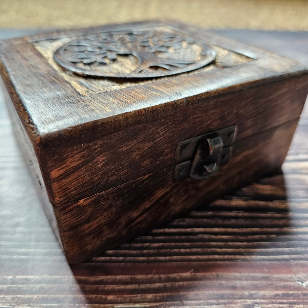 A close up of a square wooden box with a carved Tree of Life design on the lid, placed on a wooden surface.