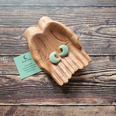 Two green aventurine crescent moon shaped crystals displayed in a pair of wooden hands on a wooden surface. 