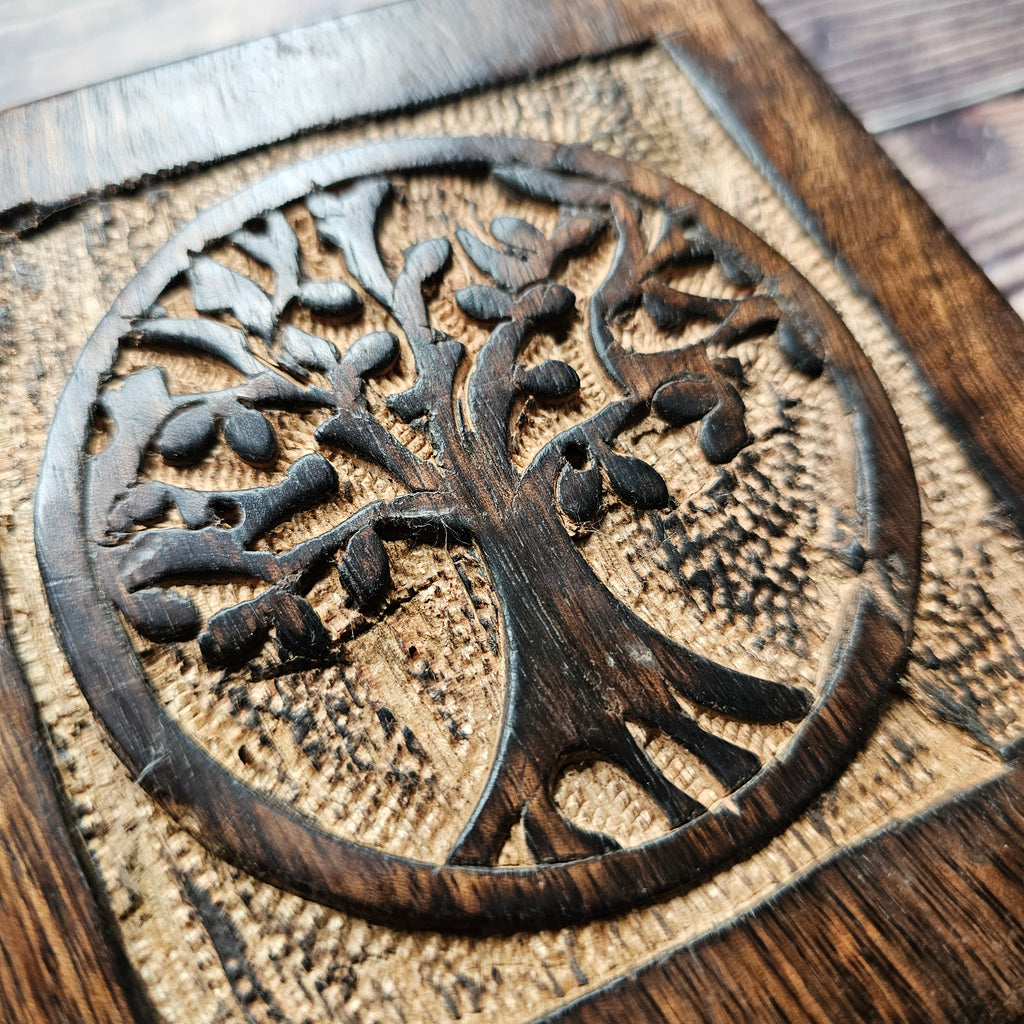 A close up of a square wooden box with a carved Tree of Life design on the lid, placed on a wooden surface.