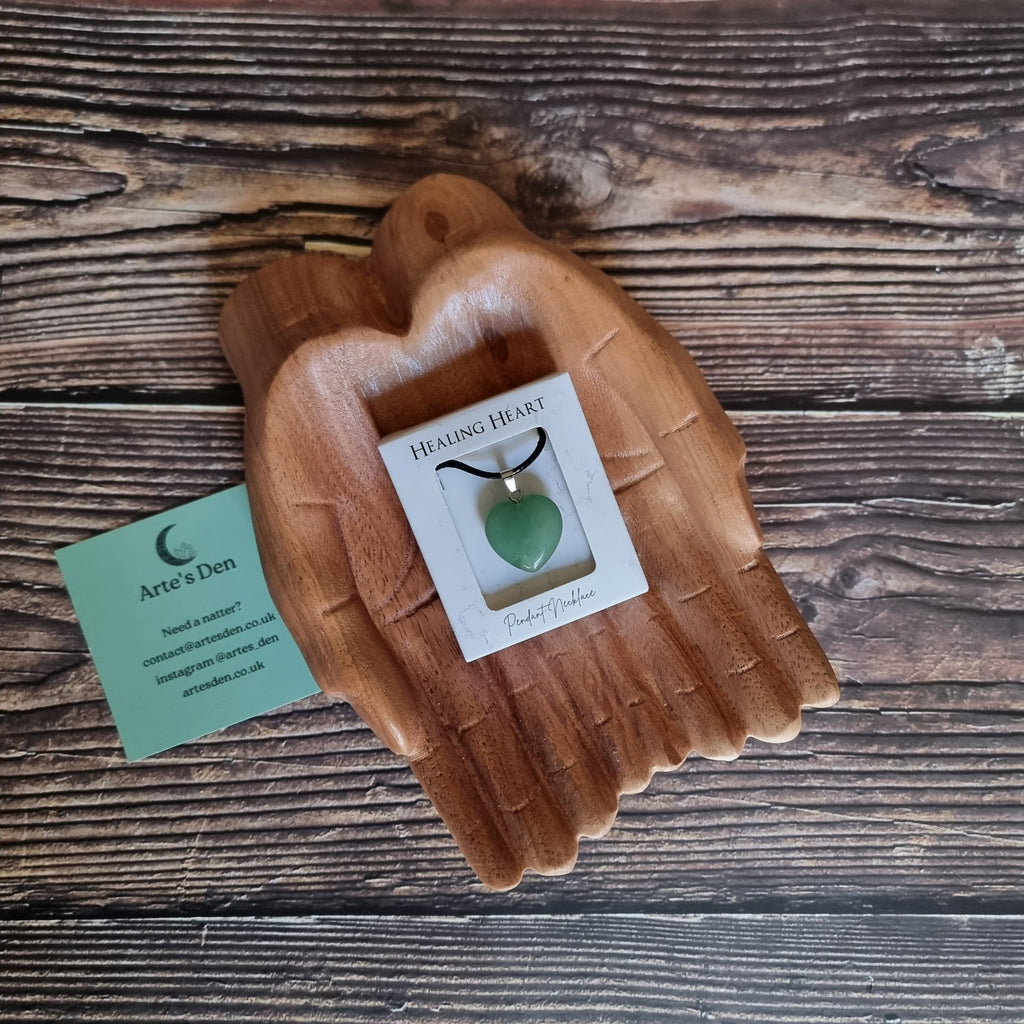 A green crystal heart on a black cord necklace inside a white display box, displayed on a pair of wooden hands