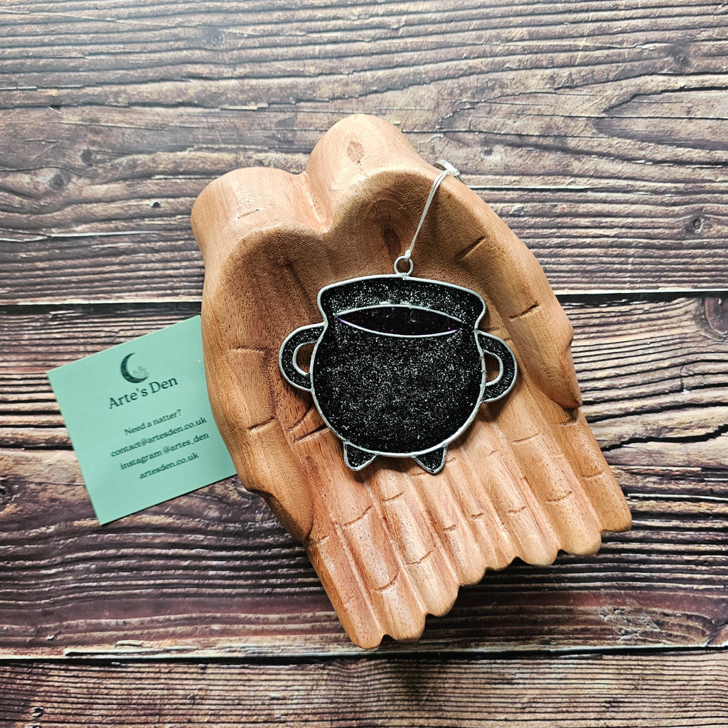A black sparkly cauldron suncatcher on a pair of wooden hands. 