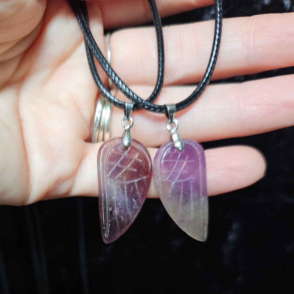 Two angel wing-shaped crystal pendants attached to a black cord necklace, held in a person's hand.