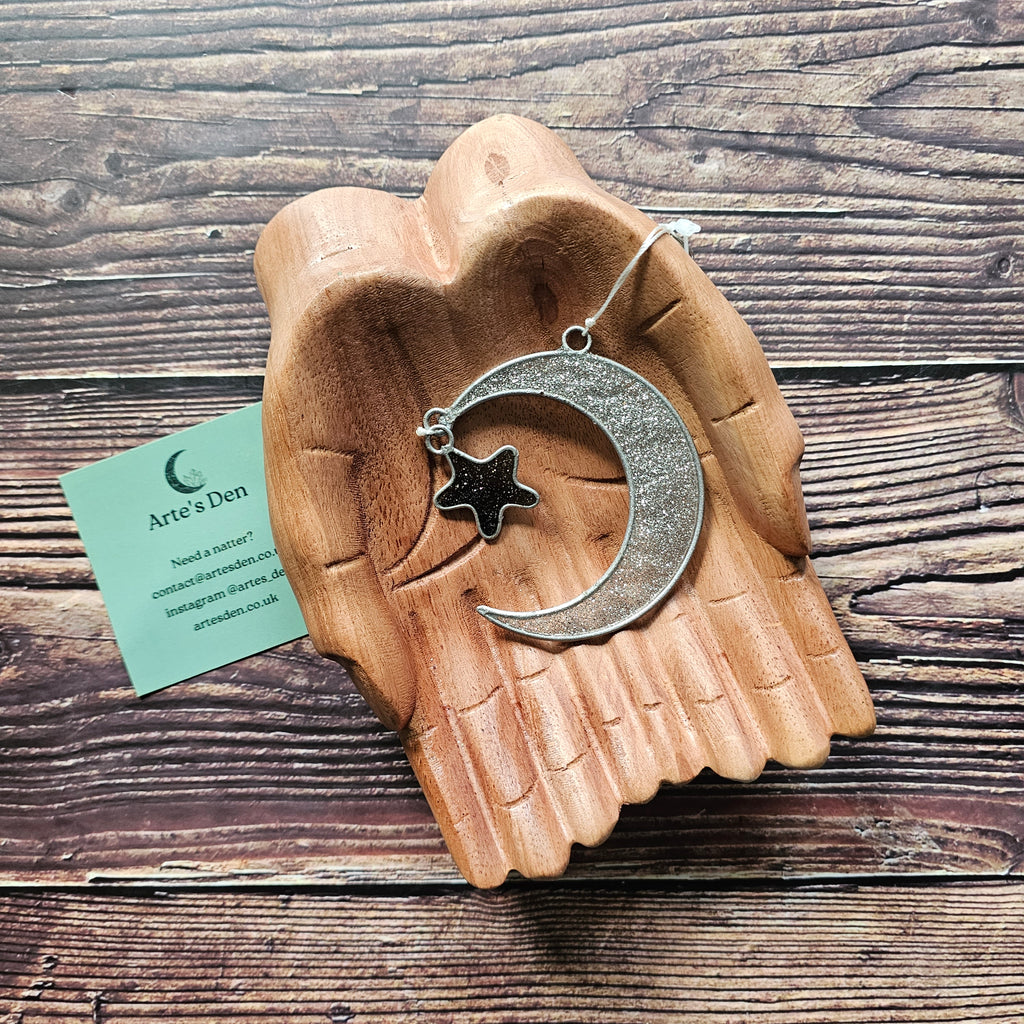 A silver sparkly crescent moon and small black star suncatcher on a pair of wooden hands. 