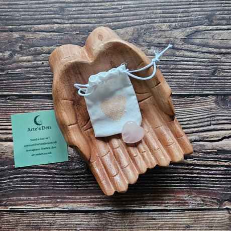 A rose quartz crystal heart stone with a small drawstring pouch, placed on a wooden pair of hands.