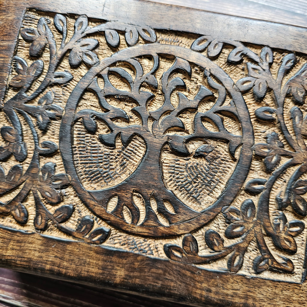 A close up of a wooden box with a carved Tree of Life design on the lid, placed on a wooden surface.