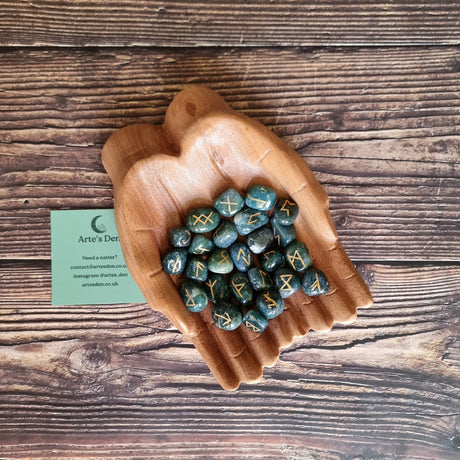 A collection of Moss Agate rune stones displayed in a pair of wooden hands, on a wooden surface with a business card labelled 'Arte's Den' on the side.