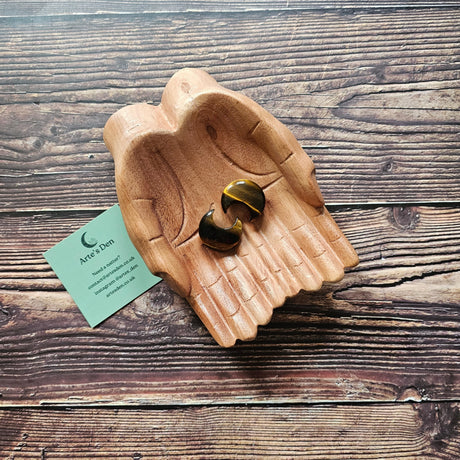 Two carved crescent moon gold-brown banded Tiger's Eye crystals placed on a pair of wooden hands against a wooden background with a card reading 'Arte's Den'.