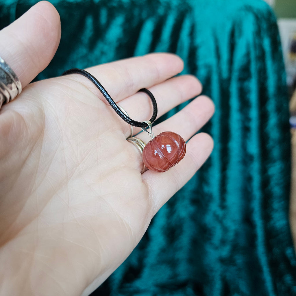 A pumpkin-shaped carnelian crystal pendant attached to a black cord necklace, held in someone's hand