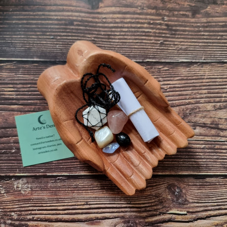A collection of crystals, a necklace and a scroll of paper displayed in a pair of wooden hands, with a business card that reads 'Arte's Den'.