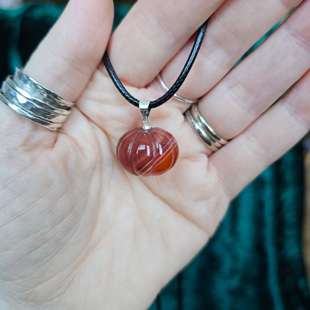 A pumpkin-shaped carnelian crystal pendant attached to a black cord necklace, held in someone's hand