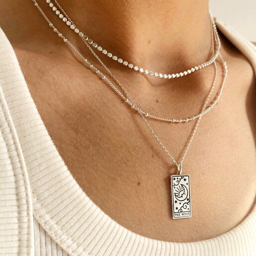 A close up of a person wearing layered necklaces including a silver tarot necklace with a pendant featuring a rectangular design, the 'Moon' card from tarot.